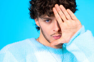 Promotional photo for "Delusional" which sees a headshot of Scott the Pisces in the photo studio, wearing a baby blue jumper with one hand up against his face with his palm out to the camera.
