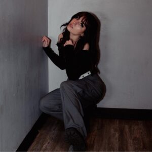 Promotional image for "Fading" which sees Calliope Wren crouching in a corner, wearing dark grey trousers and a black long-sleeved top, with her make-up looking gorgeous.
