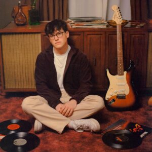 Official single cover artwork for "hope u feel ok" which sees Rob Eberle sitting crossed-legged on the floor with a guitar to his right leaning against a cabinet, and vinyl records on the floor around him. He is wearing beige trousers, white trainers, a brown jacket, and a white t-shirt.