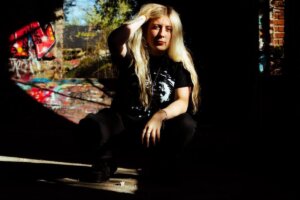 Madison Arruda wearing black clothes with a hand through her blonde hair, sitting outside with graffiti behind her.