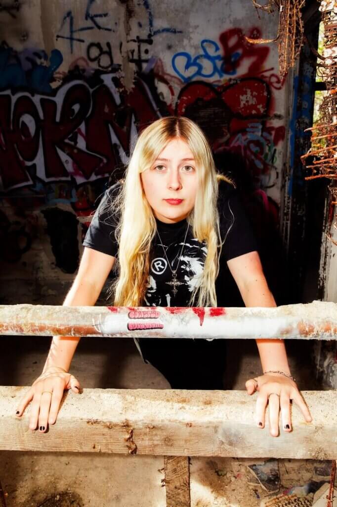 Madison Arruda leaning forward with her hands in front of her on a small wall with a metal railing. She's wearing black clothes and there's graffiti behind her.