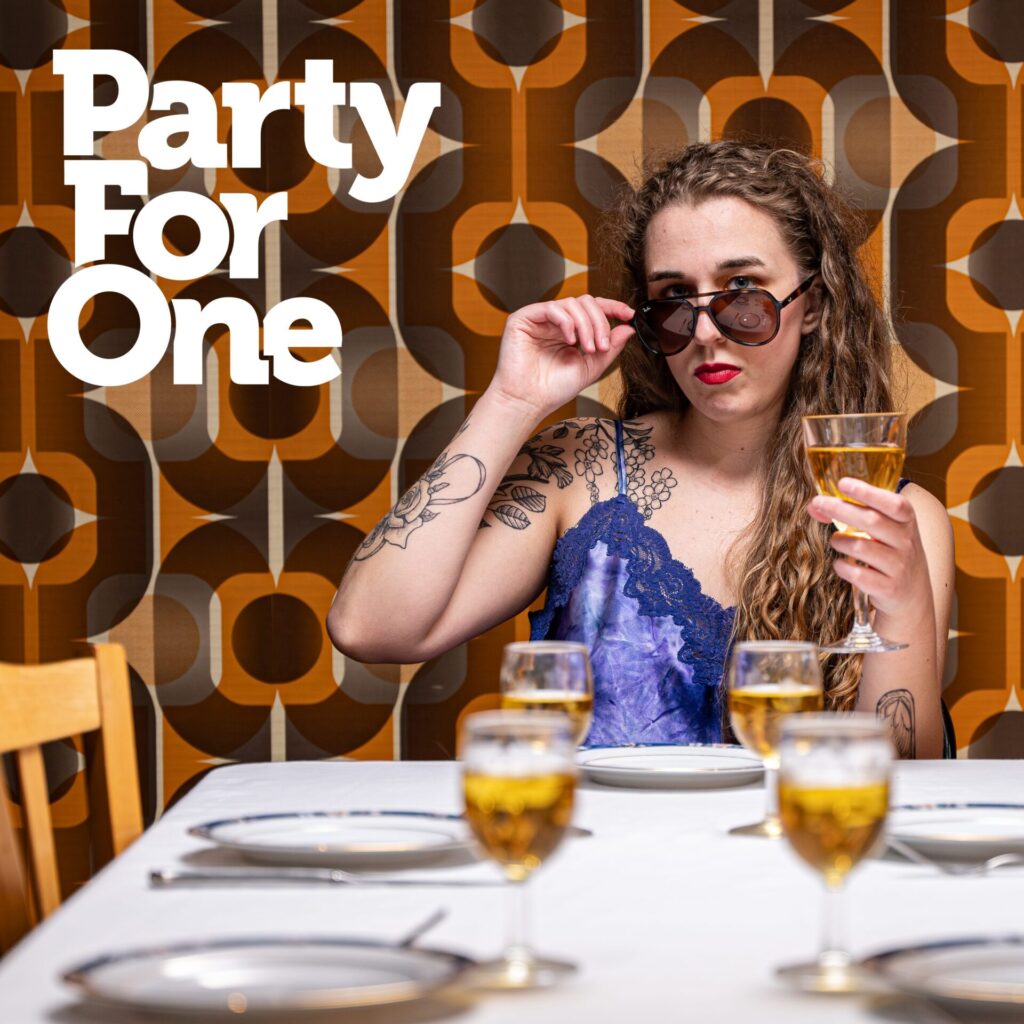Official single cover artwork for "Party For One" which sees Leanne Gallati seated at a dining room table in a blue dress, wearing some thick sunglasses which he is peering over the top of, in judgement.