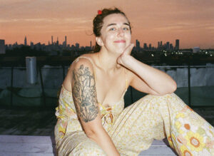 Leanne Gallati wearing beige clothes while sitting on top of a metal beam on top of a roof, overlooking the city skyline.