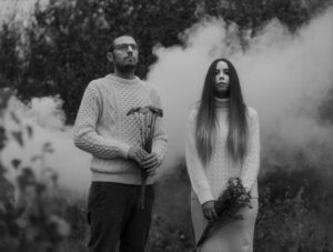 Black and white photo of the duo Remember Summer in a field with Paddy Conn on the left wearing a jumper and holding some flowers out in front of him, and Angelina Dove on the right wearing a long-sleeved dress and holding a bouquet of flowers in her hands holding them down in front of her.