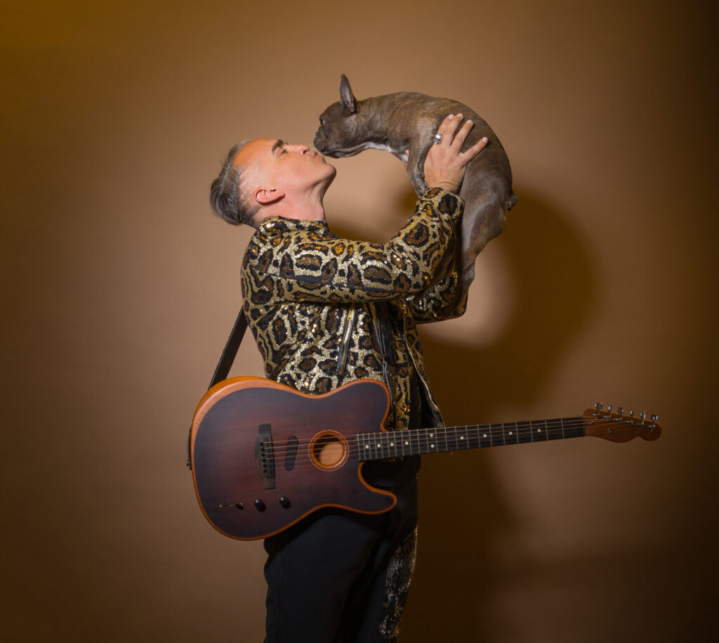 Roger Kuhn posing in front of a beige background, wearing a leopard-print shirt and black trousers with a guitar facing us with the strap over his shoulder while he faces to the right, holding a small dog in his hands, held in the air as they kiss.