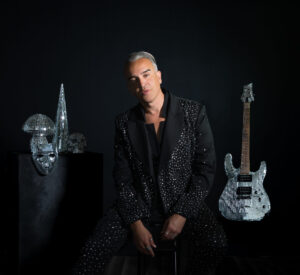Roger Kuhn posing for a photo, dressed in black with a black background, while a silver glittery guitar is to his left and a matching mask to his right.