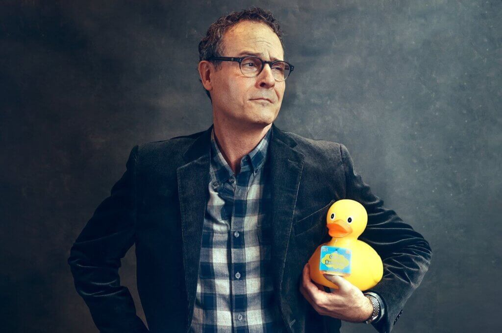 Professional photo of Keith A. Getchell standing against a photographer's typical backdrop, holding a rubber duck under his left arm.