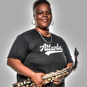 Official professional photo of Brandon-Nicole wearing a black t-shirt and smiling while holding her saxophone in fron to her.