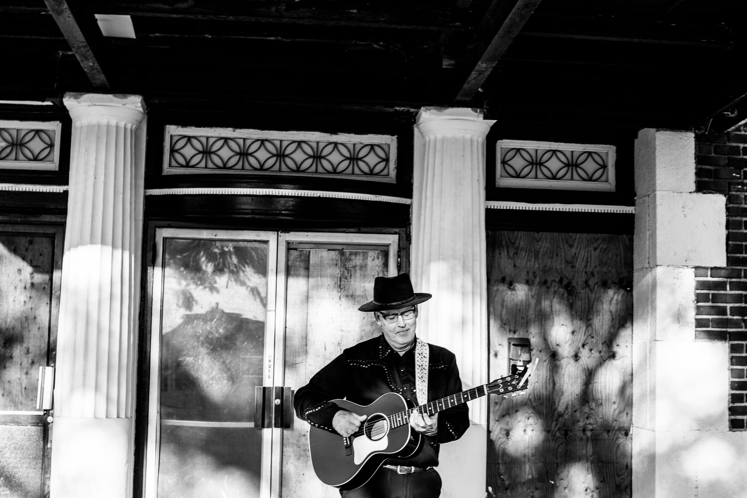 Black and white photo of Fran O'Connell performing as The FrannyO Show on the streets, outside a store, with a guitar around him and a hat. It looks like he's busking.