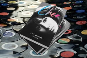 Three books on top of each other with the cover of "Cher: The Memoir, Part One", while the background is a collection of vinyl records all strewn on the floor together.
