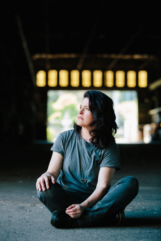 Stephanie Schneidman posing in the Zidell Shipyards wearing a loose-fitted grey t-shirt sitting on the floor looking to the left.