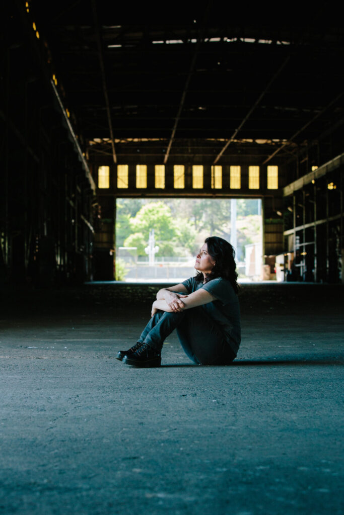 Stephanie Schneidman posing in the Zidell Shipyards wearing a loose-fitted grey t-shirt sitting on the floor with her knees to her chest, looking to the left.