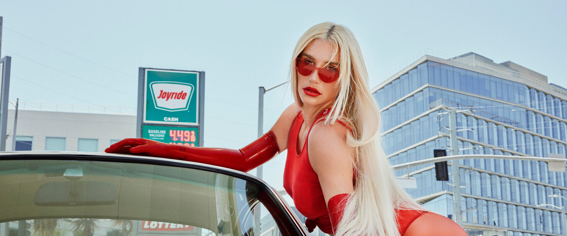 Kesha wearing a red outfit, leaning sexually on the side of a car with both hands on the vehicle, whilst looking at the camera. Her long blonde hair falls down her body and she's wearing bright red sunglasses.