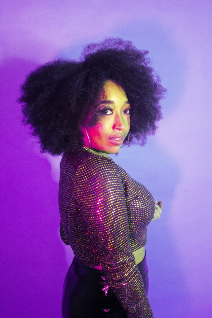 Professional photo of Cylla Senii, posing in front of a purple background, looking over one shoulder at the camera and wearing a deep purple outfit.