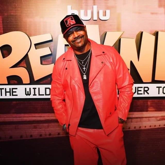 P. Frank Williams at a Hulu premiere wearing a red jacket, matching pants, a black top and a cap, His hands are in his pockets in front of a Hulu TV series logo.