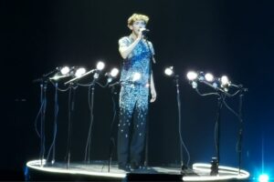 Troye Sivan performing on stage at the Utilita Arena Birmingham on his Something To Give Each Other Tour, where he's singing into a microphone, wearing a coordinated sequin blue-to-black outfit, surrounded in a circle of lights that look like microphones.