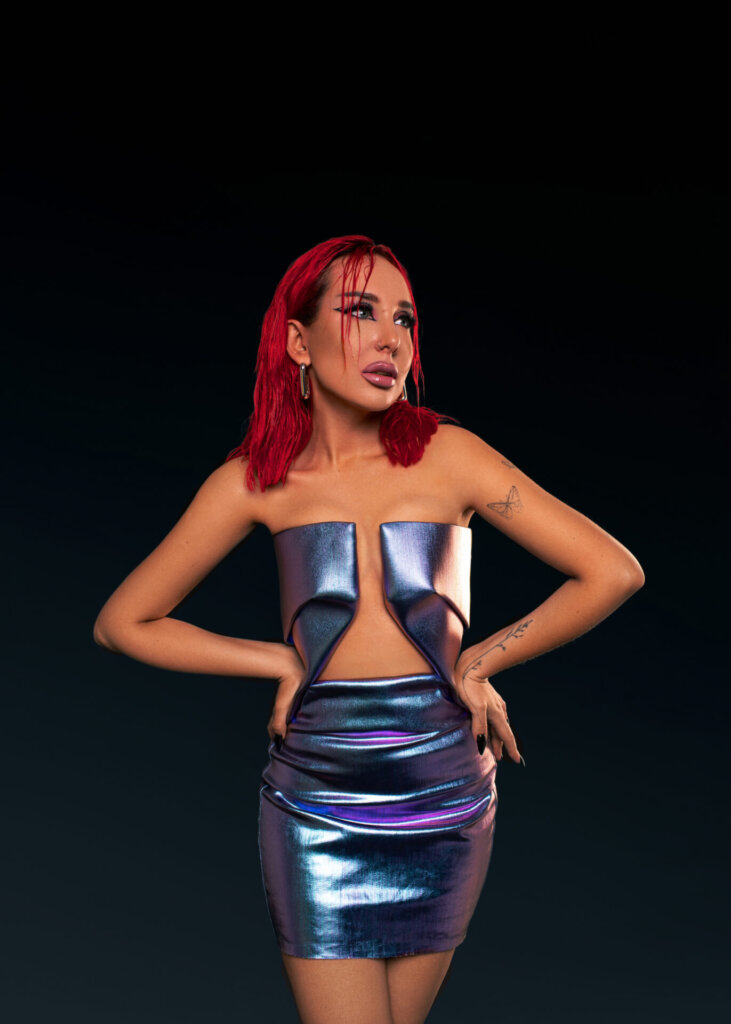 A portrait professional photo of SHADU posing in promotion of "Stillness" where she has her hands on her hips, with a metallic skirt and top which contrasts well with her bright red hair.
