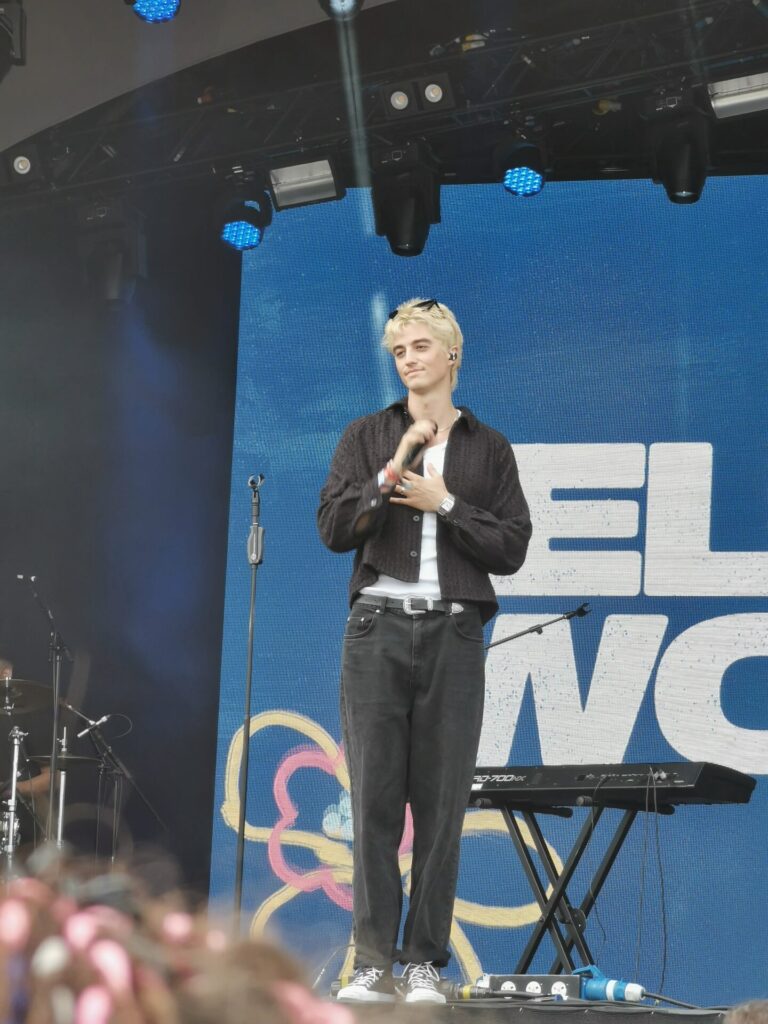 Elijah Woods performing on the Rainbow Stage at the Stray Kids-headlined BST Hyde Park. He's wearing dark jeans, a vest top, a loose overshirt, and some shades which he has on top of his head, contrasting with his blonde hair.