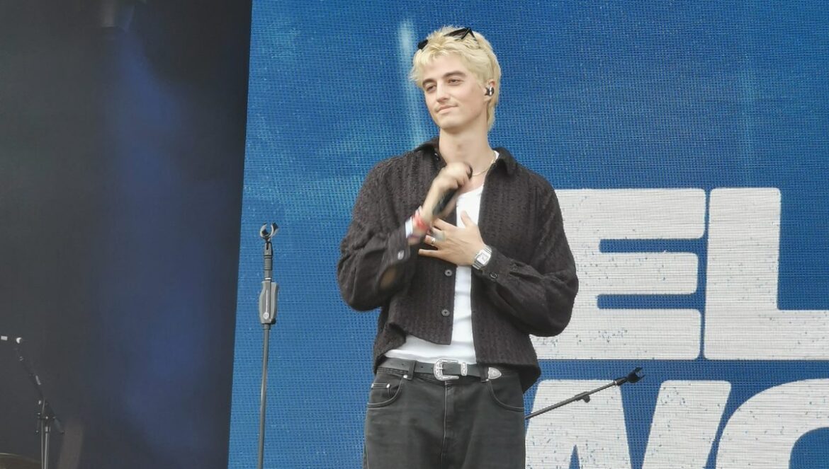 Elijah Woods performing on the Rainbow Stage at the Stray Kids-headlined BST Hyde Park. He's wearing dark jeans, a vest top, a loose overshirt, and some shades which he has on top of his head, contrasting with his blonde hair.