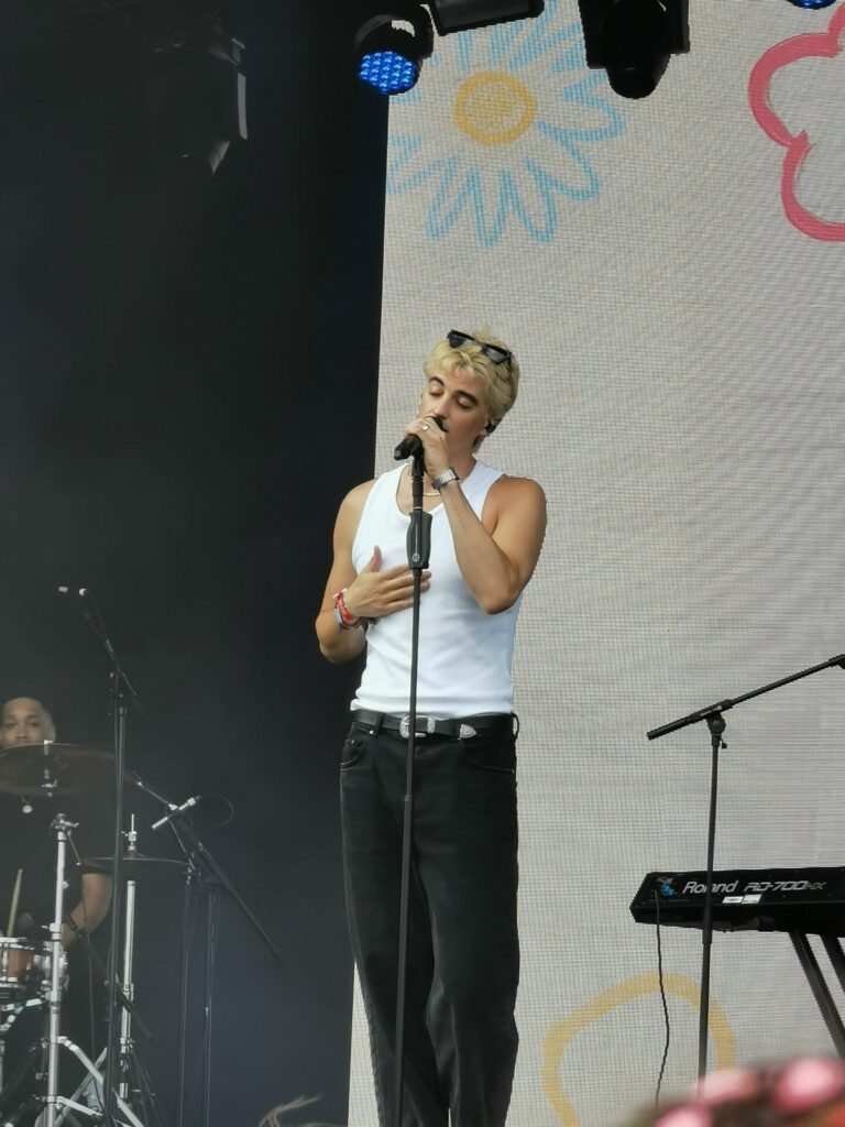 Elijah Woods performing on the Rainbow Stage at the Stray Kids-headlined BST Hyde Park. He's wearing dark jeans and a vest top having taken off his overshirt, while his shades are on top of his head, contrasting with his blonde hair. He is singing into a mic which is attached to a mic stand.