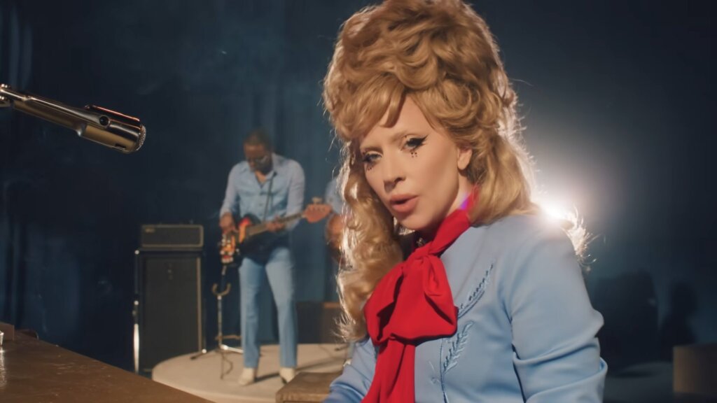 Still from the "Die With a Smile" music video which sees Lady Gaga singing to the camera where she's sitting at a piano that has a microphone stand. She's wearing a blue dress with a red thin scarf around her neck that is tied in a bow.