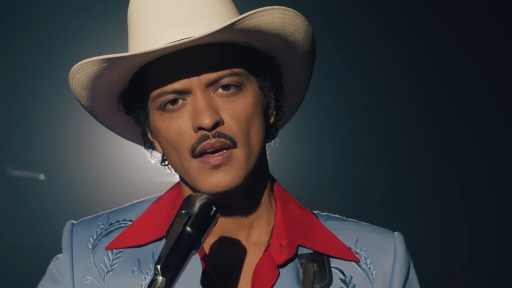 Still from "Die With a Smile" music video which sees a headshot of Bruno Mars singing into a microphone with his cowboy hat and blue suit with the red shirt collar contrasting brilliantly.