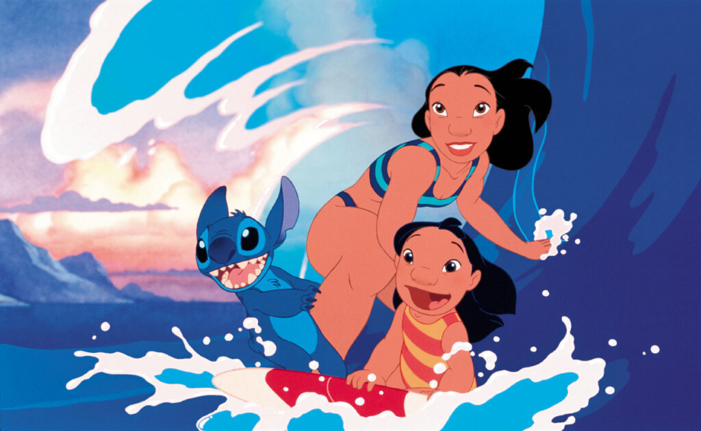 A still from the 2002 Disney "Lilo & Stitch" film which sees Lilo, Stitch, and Nani surfing on a surfboard as a huge wave curves behind them.