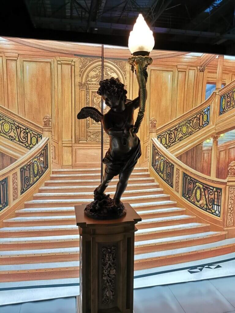 A close-up photo of a cherub statue holding a light, with the grand staircase behind.