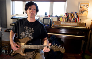 Pete Yorn performing during his Cardboard Sessions video, holding the cardboard guitar and wearing a Diana Ross band t-shirt. He is in a studio recording on the instruments.