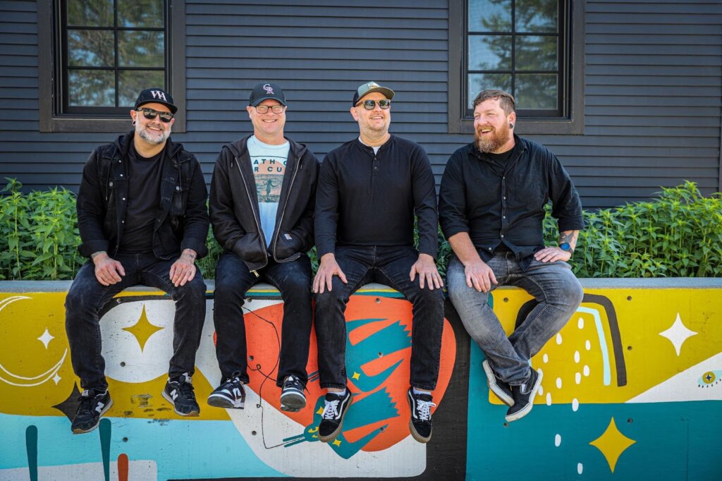 Names Without Numbers' four band members sitting on a wall that has art graffiti on it.