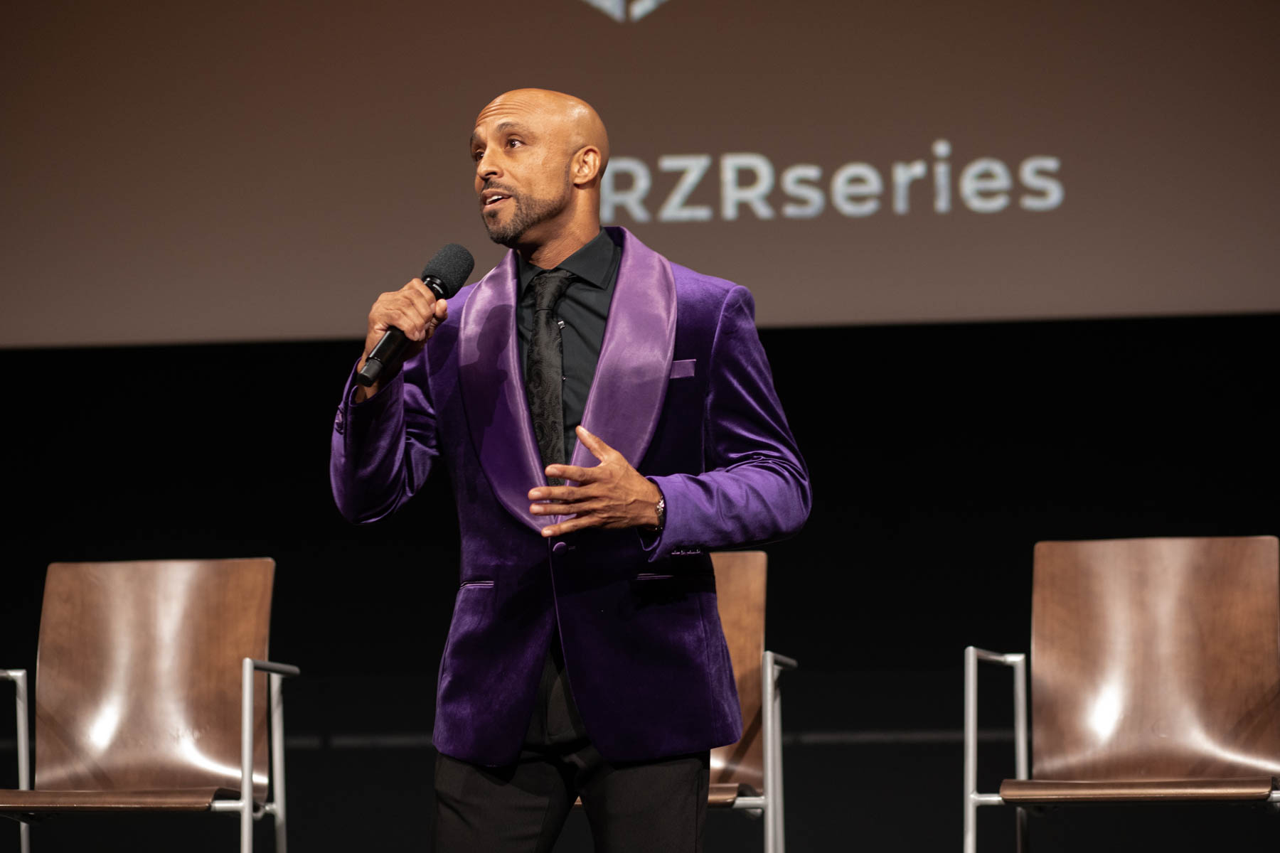 David Bianchi wearing a purple suit jacket and a black shirt, on a stage, talking into a microphone.