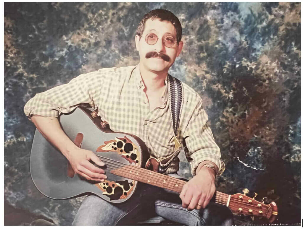 Studio photo of Denim Dan holding a guitar and wearing a white chequered shirt and jeans.