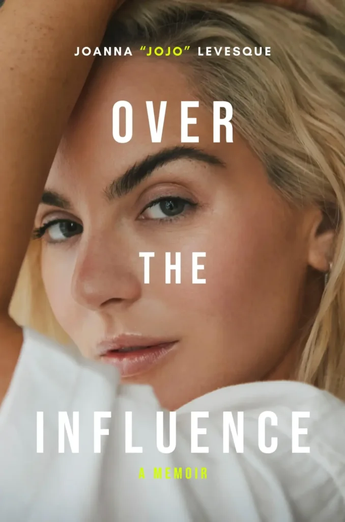 Official book cover for Joanna "JoJo" Levesque's book "Over The Influence A Memoir". It is mainly a headshot of JoJo looking over her shoulder at the camera, with her hand in her hair with her arm creating a frame around her face. Her make-up is on point and she's wearing a short-sleeve white shirt.