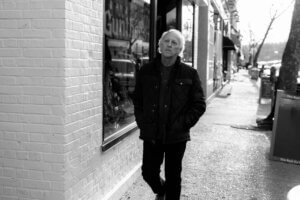 Black and white still from the "When Life's Cold" music video which sees Marshall Oakman walking down a street past a shop, with his hands in his coat pockets.