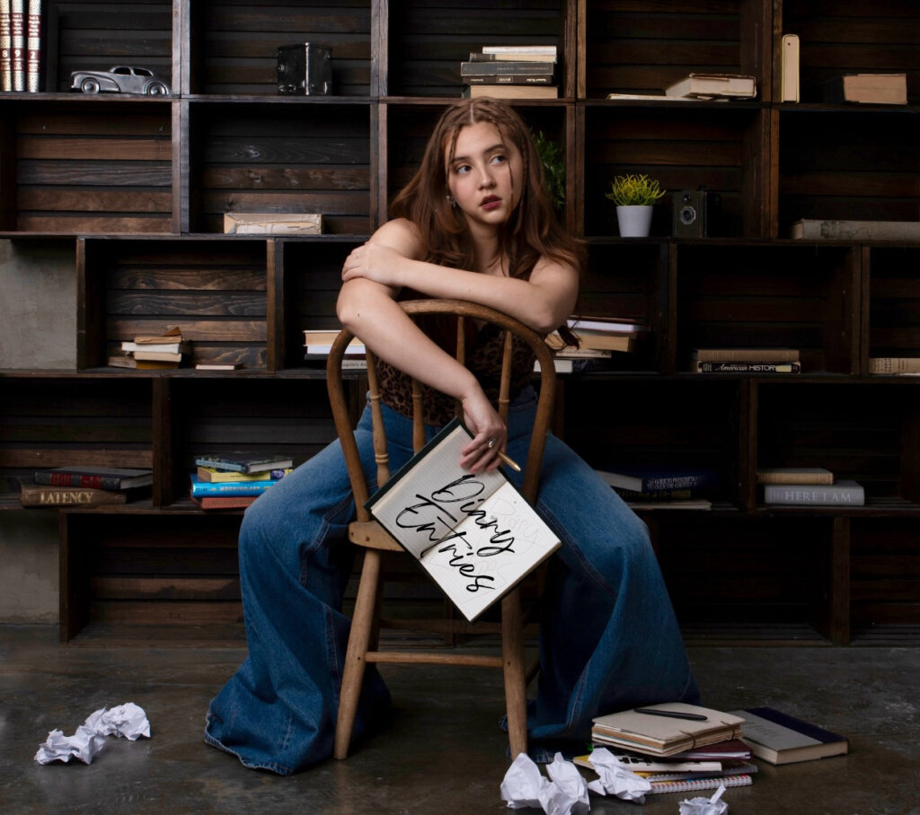 Official EP cover artwork for "Diary Entries" which sees Maya Ixta sitting backwards on a chair with a floor-to-ceiling shelfing unit behind her and paper screwed up on the floor. She's wearing a pair of jeans with a huge boot-leg flare at the bottom of them, and she's holding a notebook open that has the title written in calligraphy lettering.