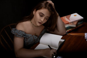 Promotional photo which sees Maya Ixta at a desk writing in a notebook as if it's her diary with her hair over one shoulder and wearing an off-the-shoulder top with a choker gold necklace paired with a necklace that falls to her chest with the letter M on it.