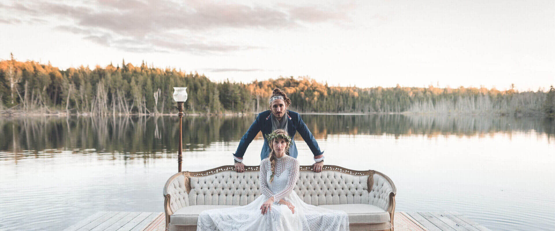 A gorgeous photo of Somewhere in Between posing on a boardwalk on a lake with Deena Robertson wearing a beautiful wedding dress, sitting on a couch with Austin Bisnow standing behind her. A forest can be seen beyond the lake.