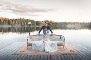 A gorgeous photo of Somewhere in Between posing on a boardwalk on a lake with Deena Robertson wearing a beautiful wedding dress, sitting on a couch with Austin Bisnow standing behind her. A forest can be seen beyond the lake.