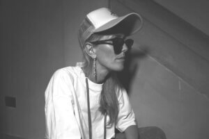 Black and white photo of Bird sitting to the side showing her right side to us, wearing a baseball cap and a white t-shirt with her hair falling over her left shoulder.