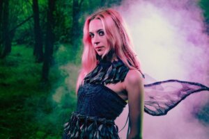 Lizzie Esau posing in a forest, wearing a black dress that highlights her pink hair, paired with fairy wings. Pink smoke is trailing behind her.