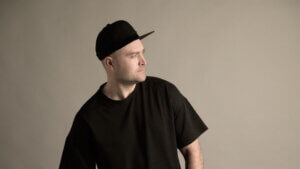 Professional photo of Seve Clain wearing a black t-shirt and a matching black snapback cap, with his head turned to the right.