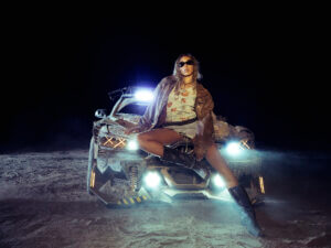 Tinashe wearing thigh-high black boots, with a grey short mini skirt, a yellow-to-green horizontal striped shirt with symmetrical cut-out circles in it, paired with a brown leather jacket and sunglasses. She's posing at night on the bonnet of a car that has its headlights on.