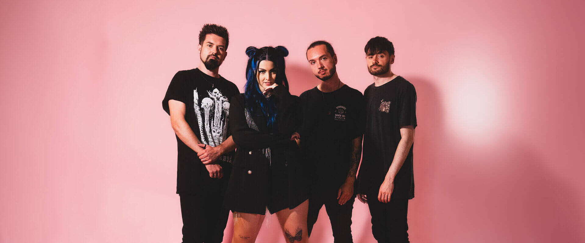 Four-piece female-fronted rock band As December Falls, dressed in black, standing together in front of a bubble-gum pink background.