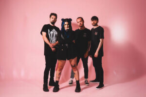 Four-piece female-fronted rock band As December Falls, dressed in black, standing together in front of a bubble-gum pink background.