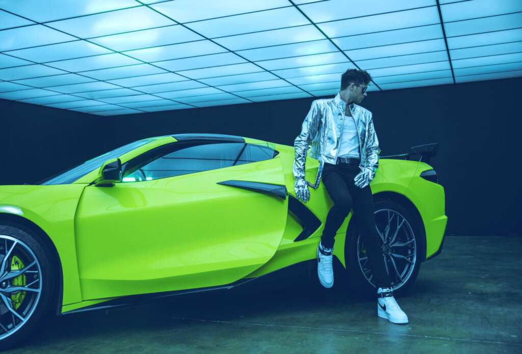 Promotional photo for the "EVEN" music video which sees Stewart Taylor leaning against a green corvette, wearing a silver jacket over a white vest, jeans, and white and black Nike trainers.