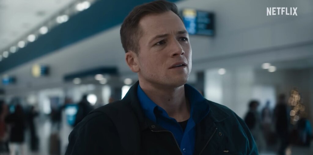 Still from Netflix's Carry-On film trailer which sees Taron Egerton looking worried as he plays TSA agent Ethan Kopek, dressed in a dark-blue coat over a bright blue uniform shirt.