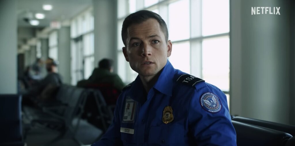 Still from Netflix's Carry-On trailer which shows Taron Egerton as Ethan Kopek in TSA agent uniform of a bright blue shirt, sitting down in the airport, chatting with the Mysterious Traveller.