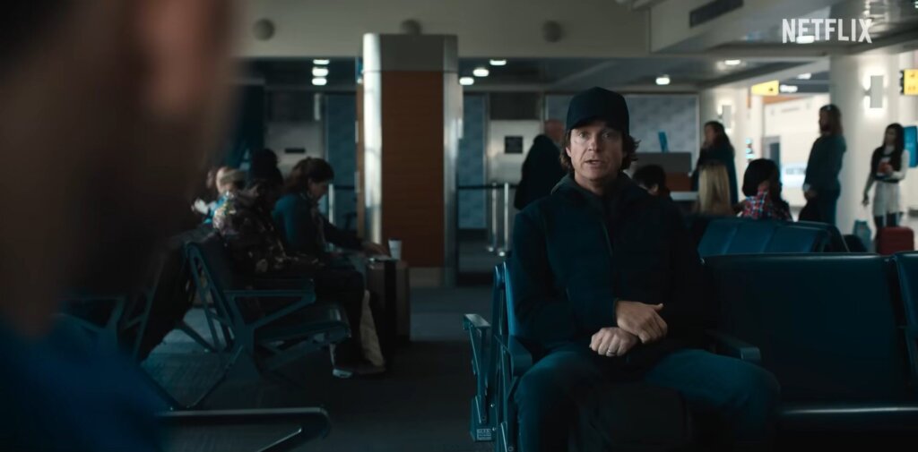 Still from Netflix's Carry-On film trailer which shows Jason Bateman as the Mysterious Traveller, dressed all in black and wearing a cap, sitting in the waiting area at the airport, opposite Taron Egerton who plays Ethan Kopek, as they discuss what's been happening at the airport.