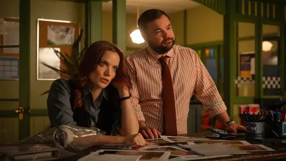 Return to Paradise still which sees Anna Samson as Mackenzie Clarke sitting at a table with Lloyd Griffith as Detective Colin Cartwright standing beside her