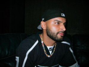 Marcel Mendoza wearing a black t-shirt with a gold chain necklace, facing slightly to the right with a black snapback cap on backwards.